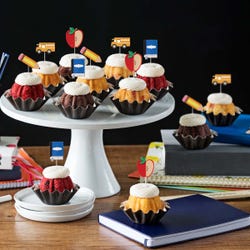 Bundtinis&reg; topped with creamy white frosting, accompanied by school-themed toppers: a sliced apple, a pencil, a notebook, and a yellow school bus.