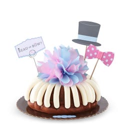 A gender reveal bundt cake with a decoration of a top hat and a pink dotted bow tie. A card reads Beau or Bow? and the center of the cake has a pink and blue bow. 