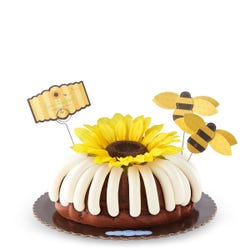 A large bundt cake with frosting. The center of the cake is topped with a large sunflower and adorned with bumblebee cutouts and a card that reads flour bouquet - Thinking of you.