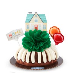 Bundt cake topped with a green pom bow and a small house decoration above it. There are two mini balloons decorating the home along with a card that reads Home Sweet Home.