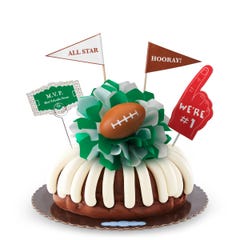 A Chocolate Chocolate Chip bundt cake with frosting, decorated with a mini football on top of a green and white bow in the center of the cake. The cake also contains a We're #1 foam finger, a flag that says All Star and a card that says MVP.