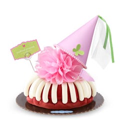 A Red Velvet bundt cake with frosting, topped with a pink bow and a princess-themed pink party hat with a butterfly on top. A card on the cake reads Prettiest Princess.