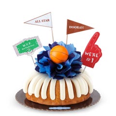 A Vanilla bundt cake with frosting, decorated with a mini basketball on top of a bow with different shades of blue in the center of the cake. The cake also contains a We're #1 foam finger, a flag that says All Star and a card that says MVP.