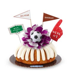A White Chocolate Raspberry bundt cake with frosting, decorated with a mini soccer ball on top of a purple and white bow in the center of the cake. The cake also contains a We're #1 foam finger, a flag that says All Star and a card that says M.V.P.