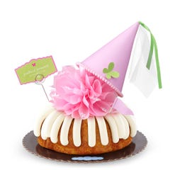 A White Chocolate Raspberry bundt cake with frosting, topped with a pink bow and a princess-themed pink party hat with a butterfly on top. A card on the cake reads Prettiest Princess.